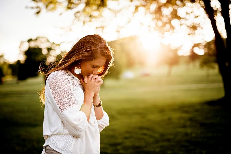 woman praying in front of a sunlit green meadow and a tree