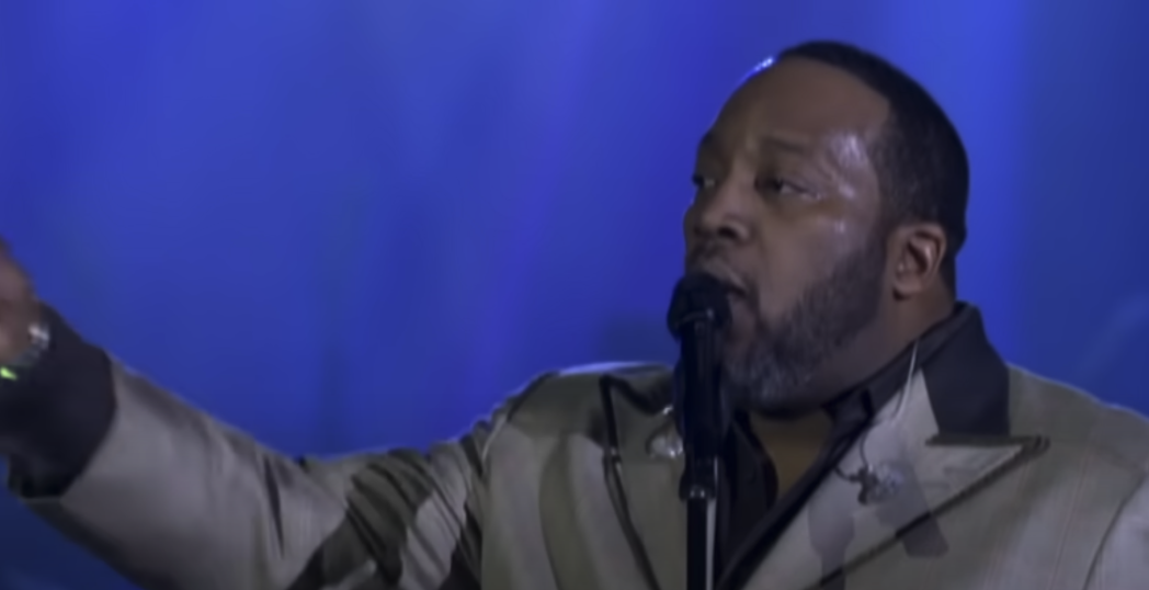 Marvin Sapp performing on a blue background while wearing a gray coat.