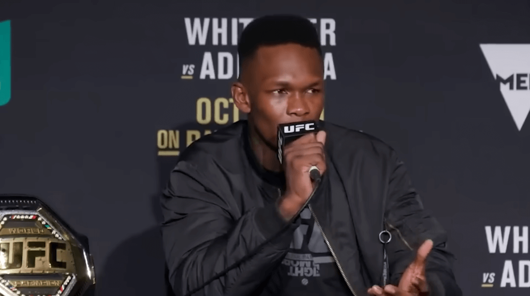 Israel Adesanya in black jacket speaking to microphone, UFC belt in the corner