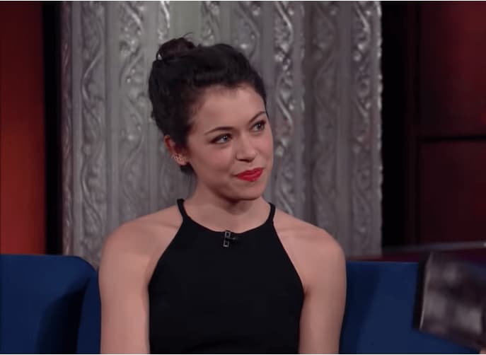 tatiana maslany in a black dress sitting on a blue sofa and looking at the side