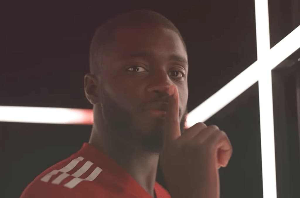 Dayot Upamecano in red and white sports clothing holds a finger near his lips, white lights behind him