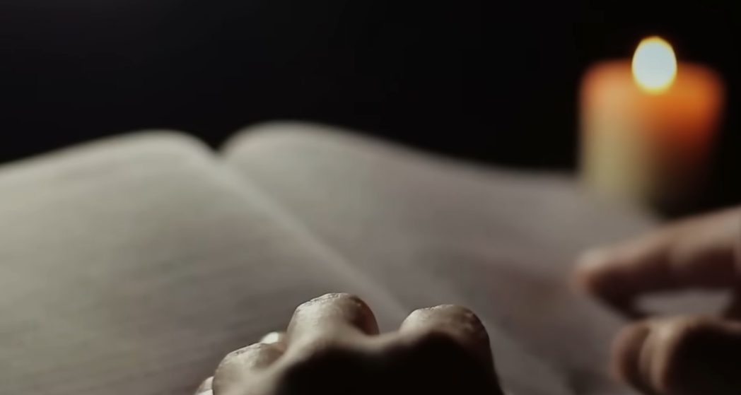 Close-up of hands on an open book with a candle in the background
