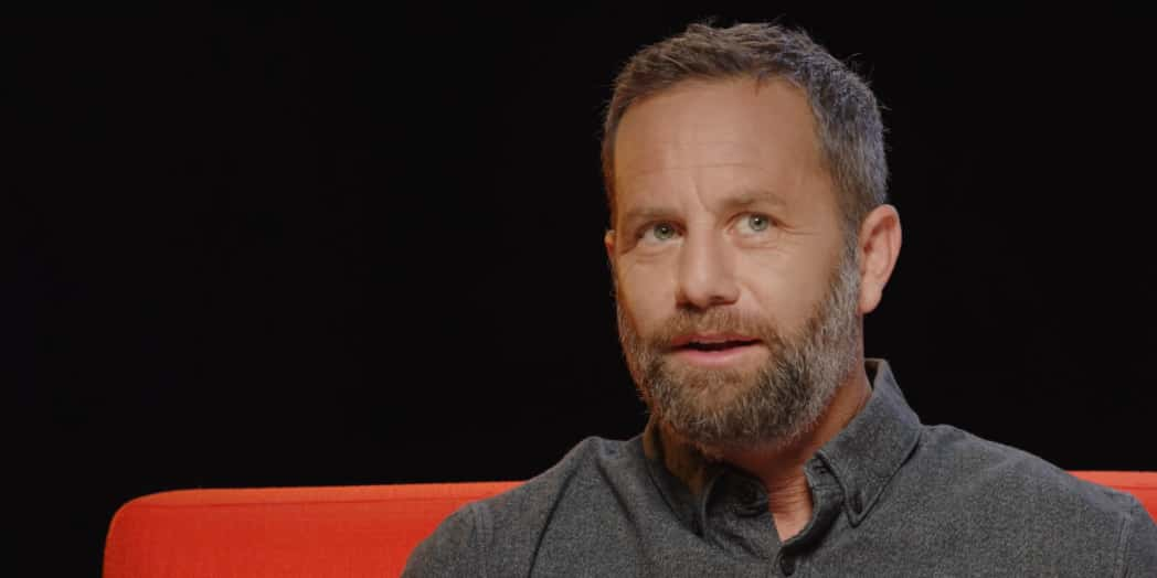 A man with a beard and a gray shirt sits, looking directly ahead