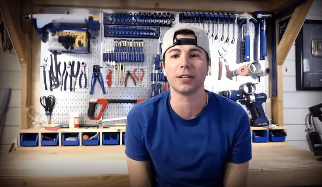 Mark Rober in a blue t-shirt and hat sitting in front of a wall with tools