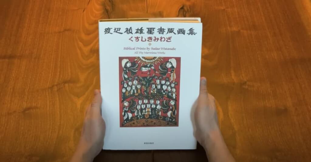 Hand holding 'Biblical Prints' book by Sadao Watanabe. White book with colorful prints and text.
