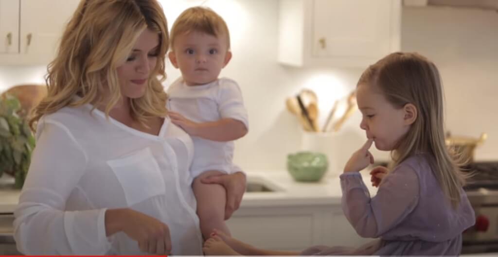 Daphne Oz cooking in the kitchen while holding her baby, with her other child sitting at the counter.