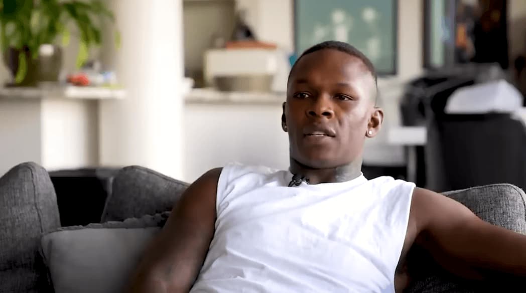 Adesanya in white t-shirt sitting on a couch in room with flowers